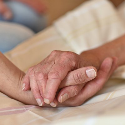 Eine Person liegt auf einem Bett. Ihre Hand wird von einer anderen Person gehalten. Auf dem Bild sind nur die Hände zu sehen.
