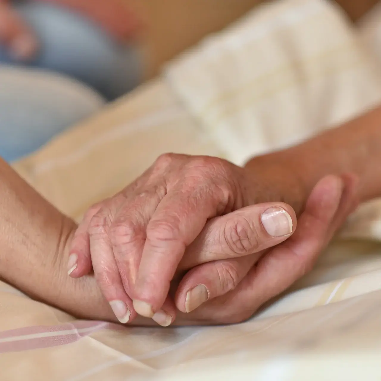 Eine Person liegt auf einem Bett. Ihre Hand wird von einer anderen Person gehalten. Auf dem Bild sind nur die Hände zu sehen.