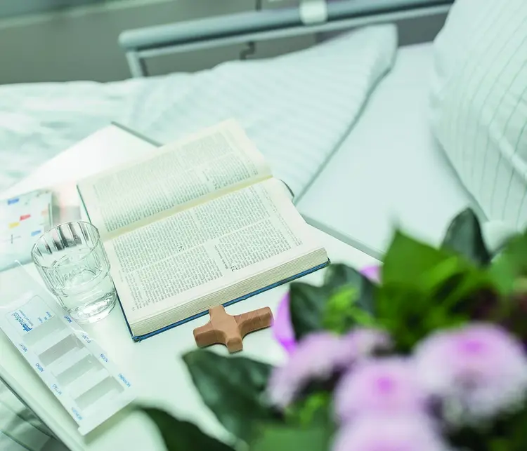 Ein Kreuz und eine aufgeschlagene Bibel liegen neben Medikamenten auf einem Krankenbett-Tisch.